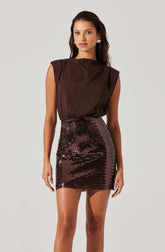 A woman is wearing a sleeveless, dark brown satin top with structured shoulders and a high neckline, paired with a high-waisted, tight-fitting mini skirt covered in shiny, dark bronze sequins.
